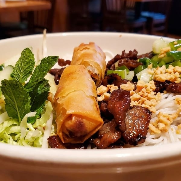 A close-up of a Vietnamese vermicelli bowl with grilled pork, spring rolls, and fresh vegetables.