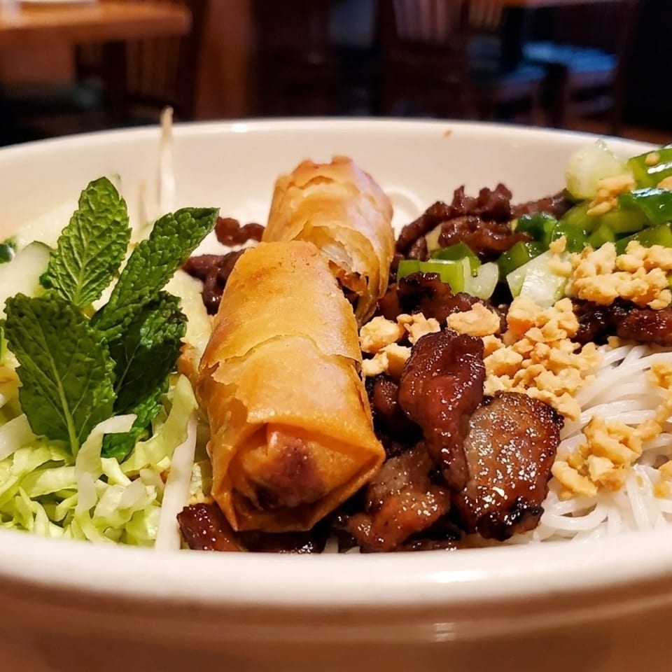A close-up of a Vietnamese vermicelli bowl with grilled pork, spring rolls, and fresh vegetables.