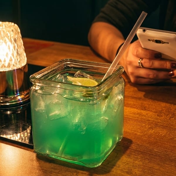 A person's hands holding a phone next to a blue cocktail and a glowing lamp on a wooden table.