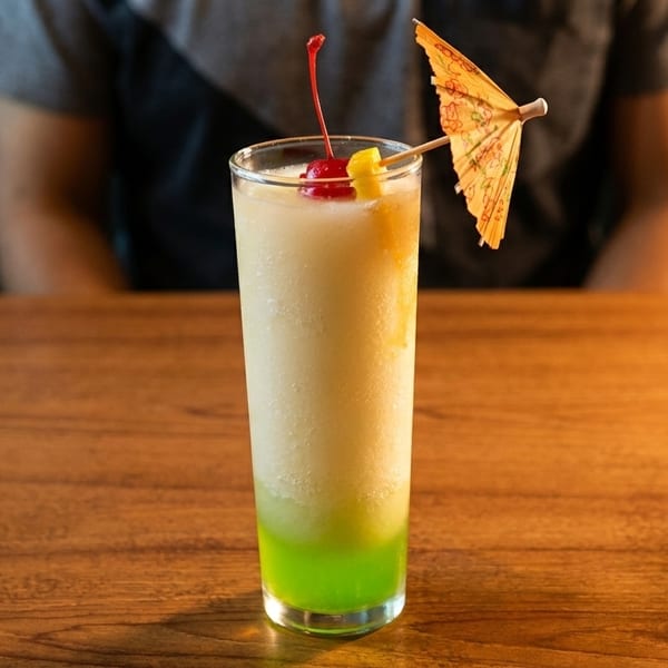 Tall tropical drink with green and white layers, cherry, pineapple, and umbrella on a wooden table.