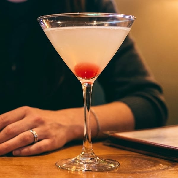 A martini glass with a light pink drink and a cherry, on a wooden table with a person's hands.