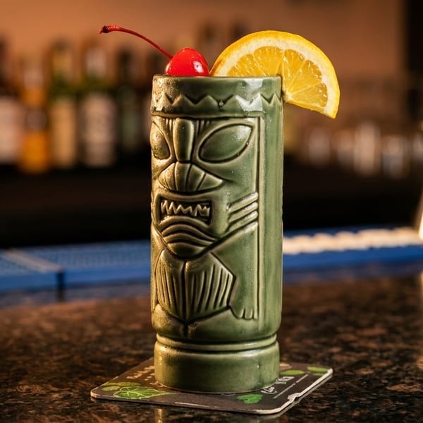 A green tiki cocktail, garnished with a cherry and orange slice, sits on a bar.